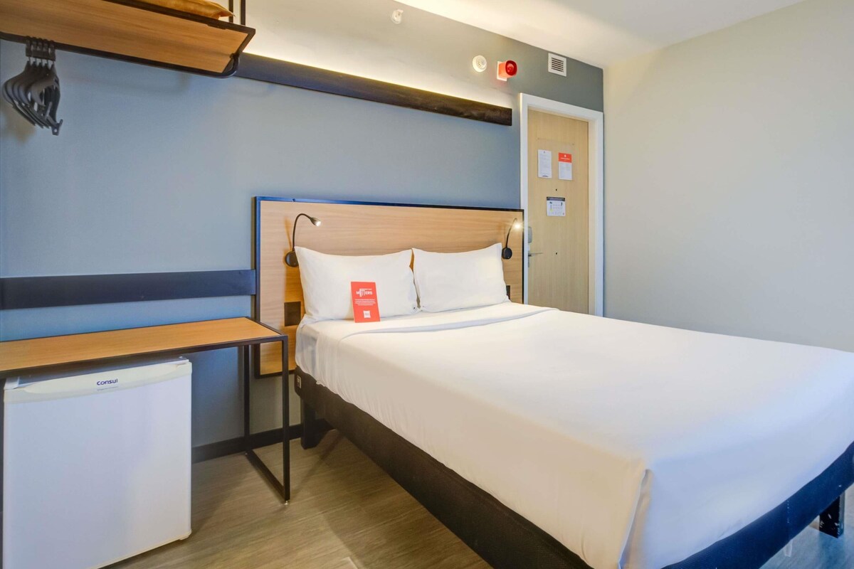 A hotel room with a bed, bedside table, and a small desk with a chair.