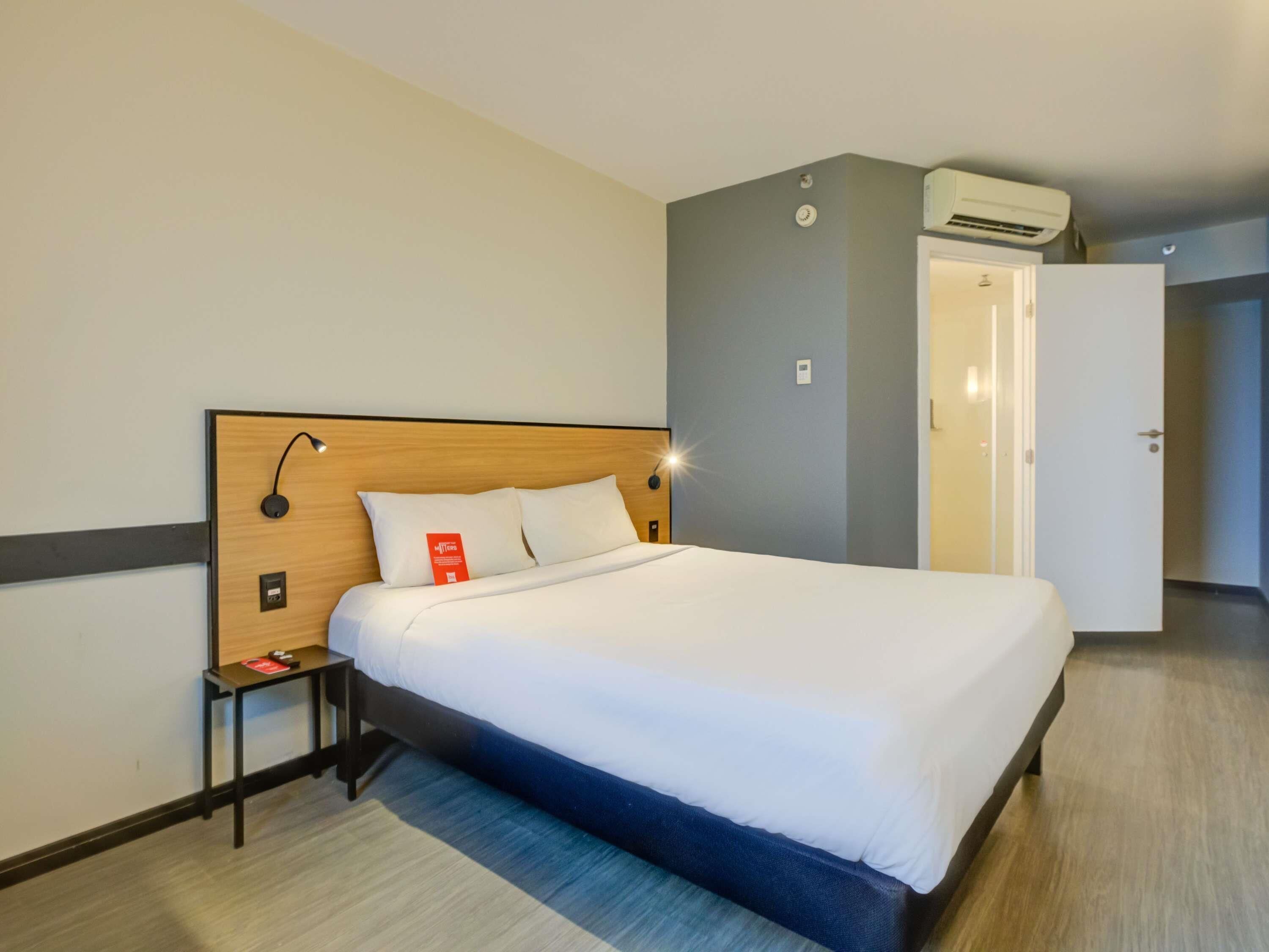 A hotel room with a bed, bedside table, lamp, and an air conditioning unit.