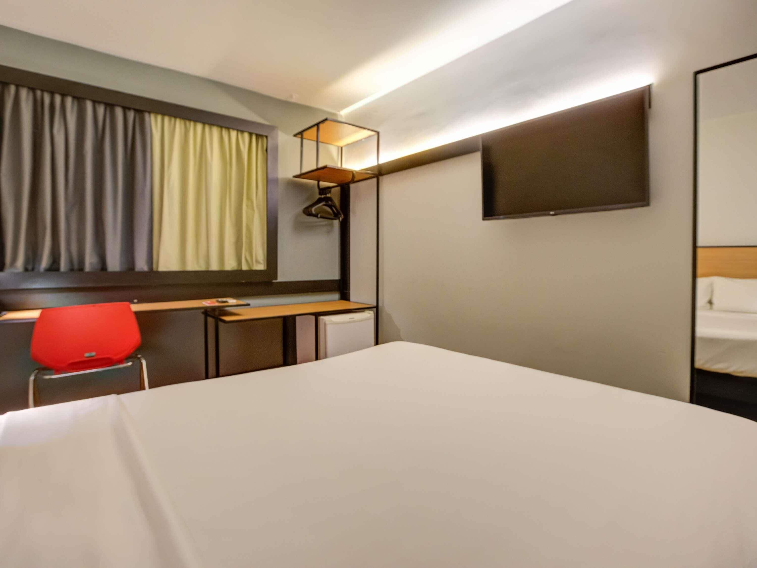 A hotel room with a bed, a desk with a chair, a TV, and a window with curtains.