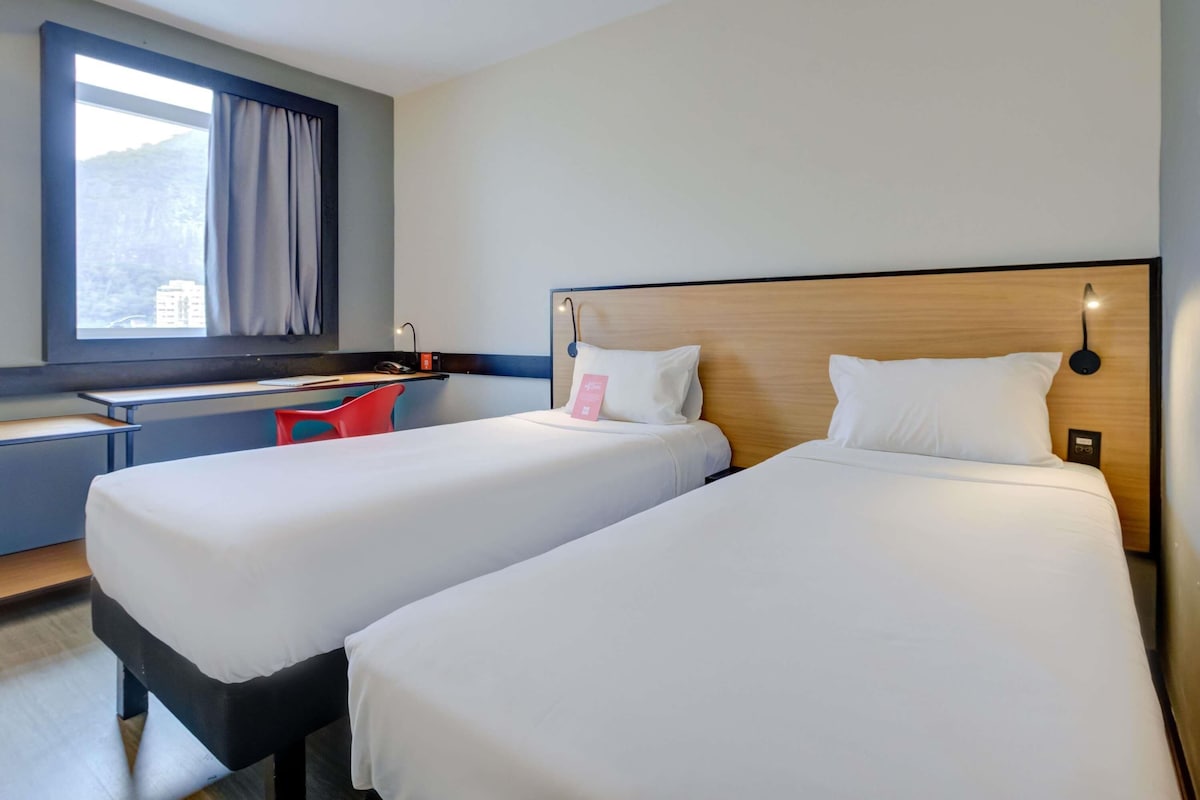 A hotel room with two single beds, a desk, a chair, and a window with curtains.