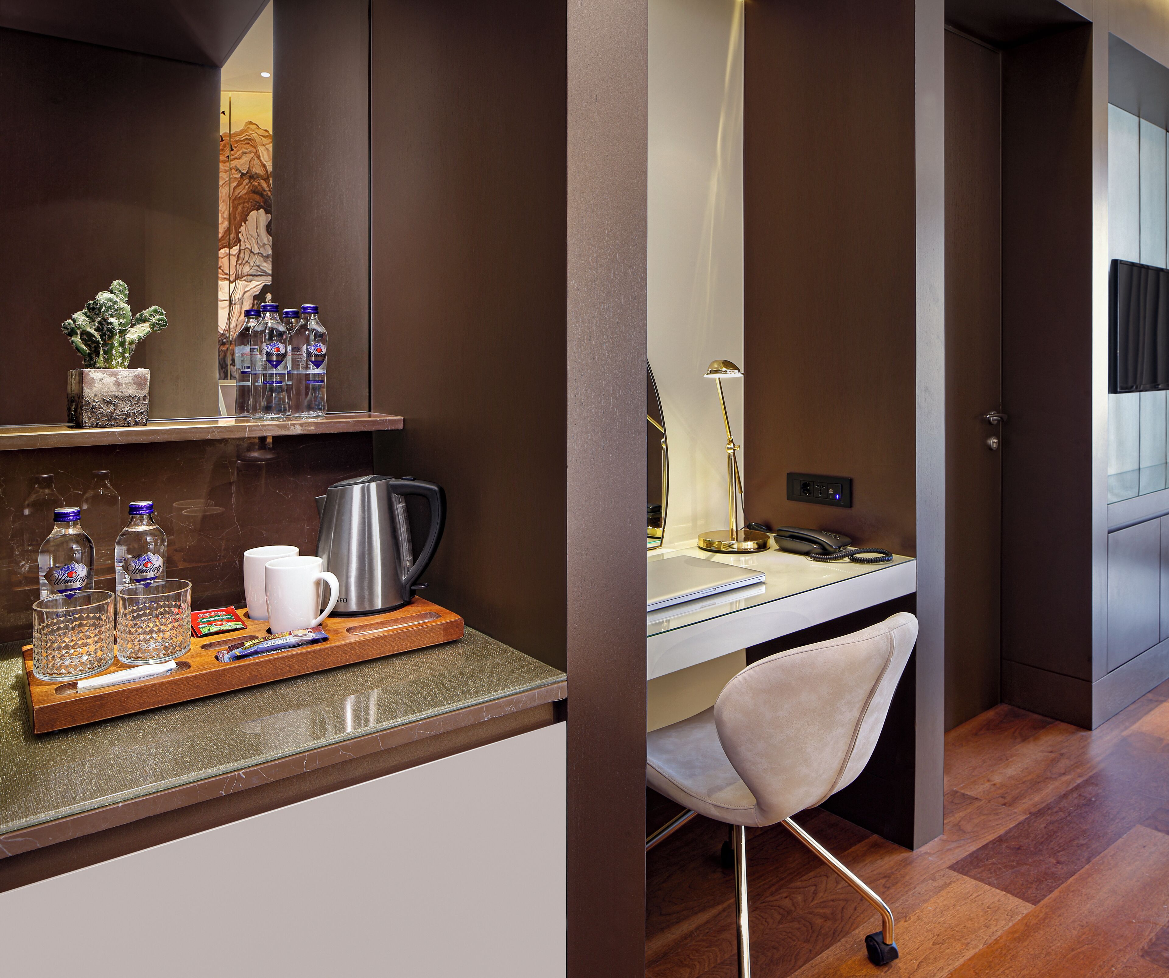 A modern hotel room with a desk, chair, coffee maker, and a shelf with bottles and a plant.