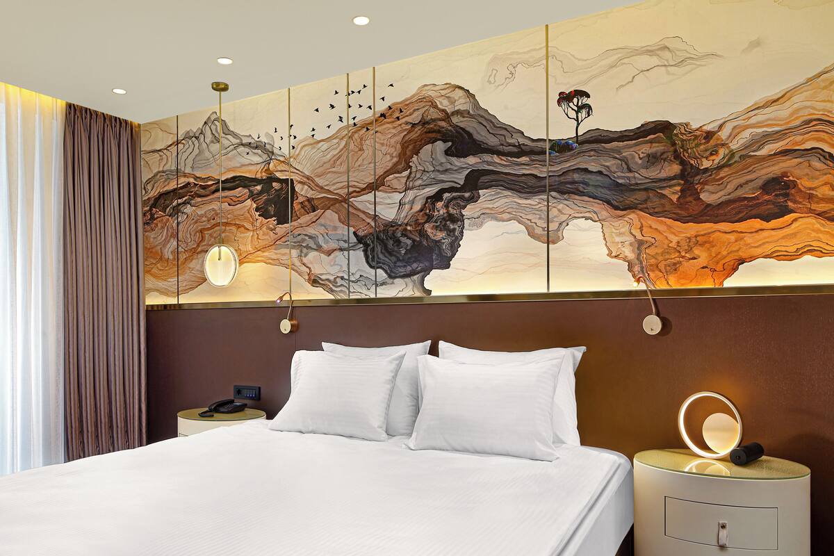 Una habitación de hotel con una cama grande, una mesita de noche con lámpara y una pared decorada con un cuadro abstracto de grandes dimensiones.