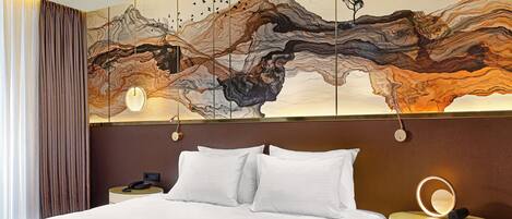 A hotel room with a large bed, a nightstand with a lamp, and a wall with a large abstract painting.