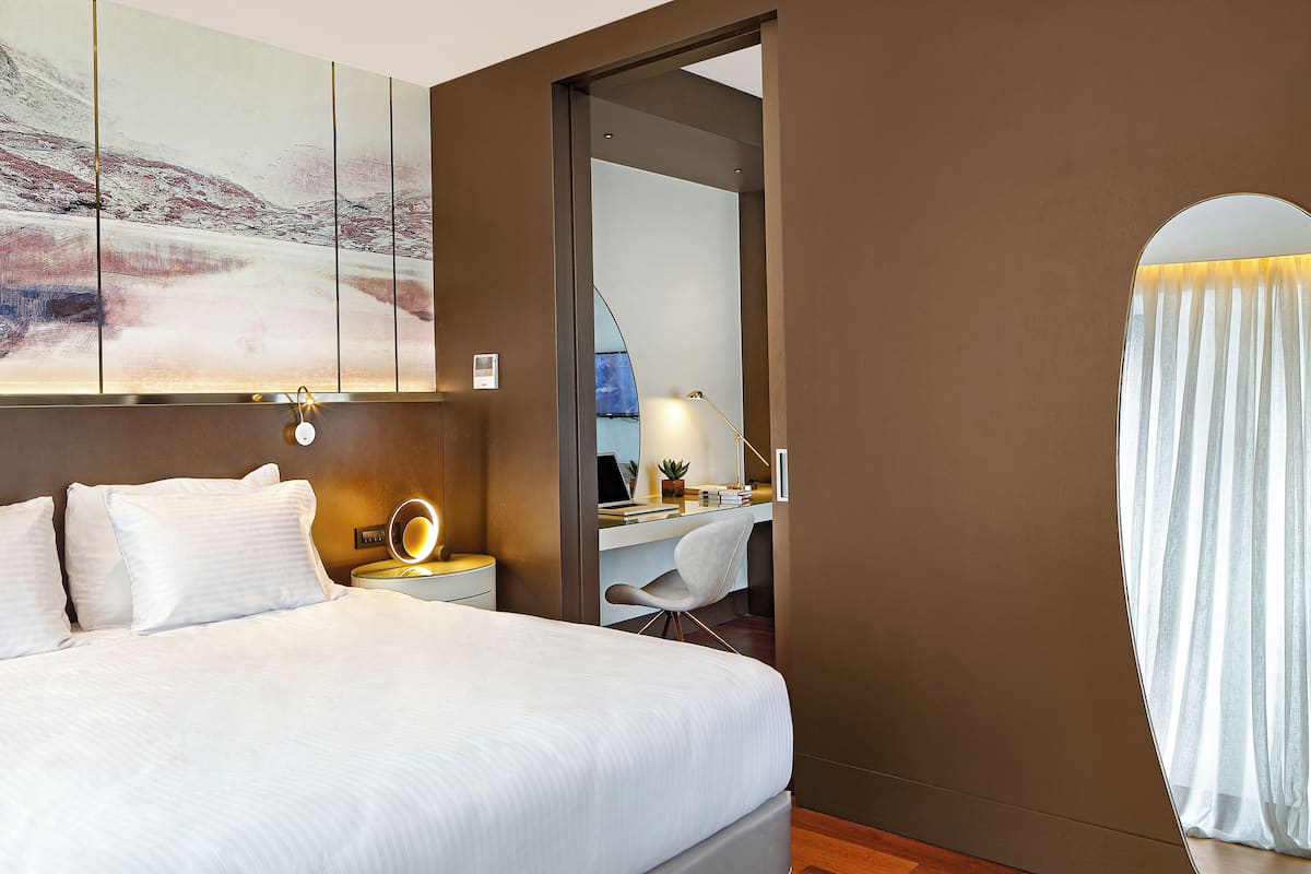 Una habitación de hotel con cama, mesita de noche, lámpara, escritorio, silla y un espejo grande.