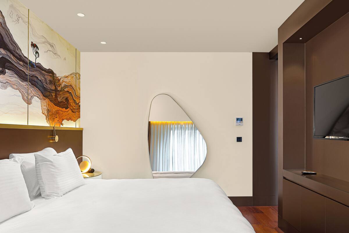 Una habitación de hotel moderna con una cama grande, un televisor, un espejo de pared original y un cuadro abstracto de grandes dimensiones.