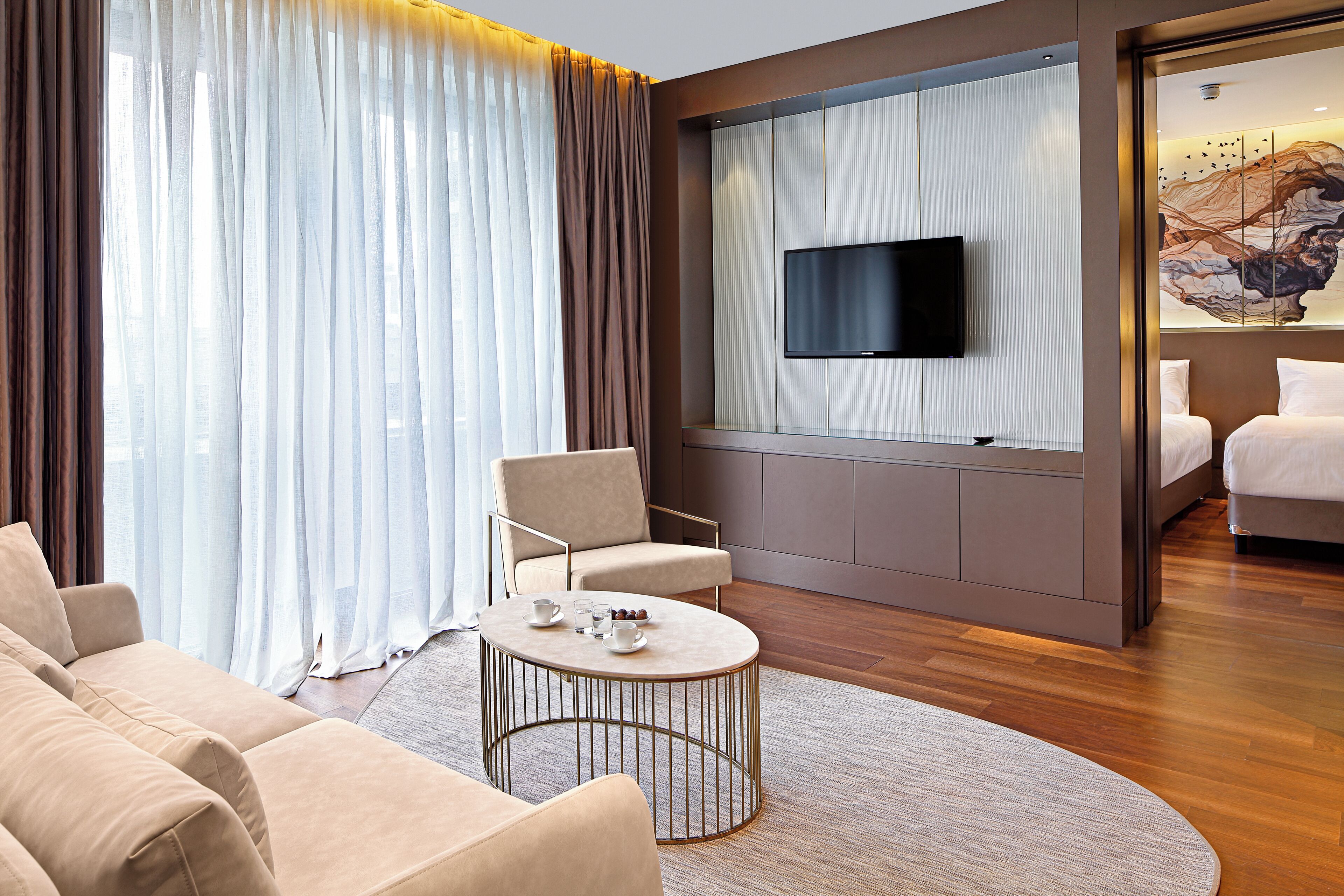 A modern hotel room with a sofa, armchair, coffee table, and a TV mounted on the wall.
