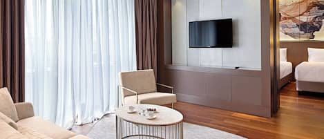 A modern hotel room with a sofa, armchair, coffee table, and a TV mounted on the wall.