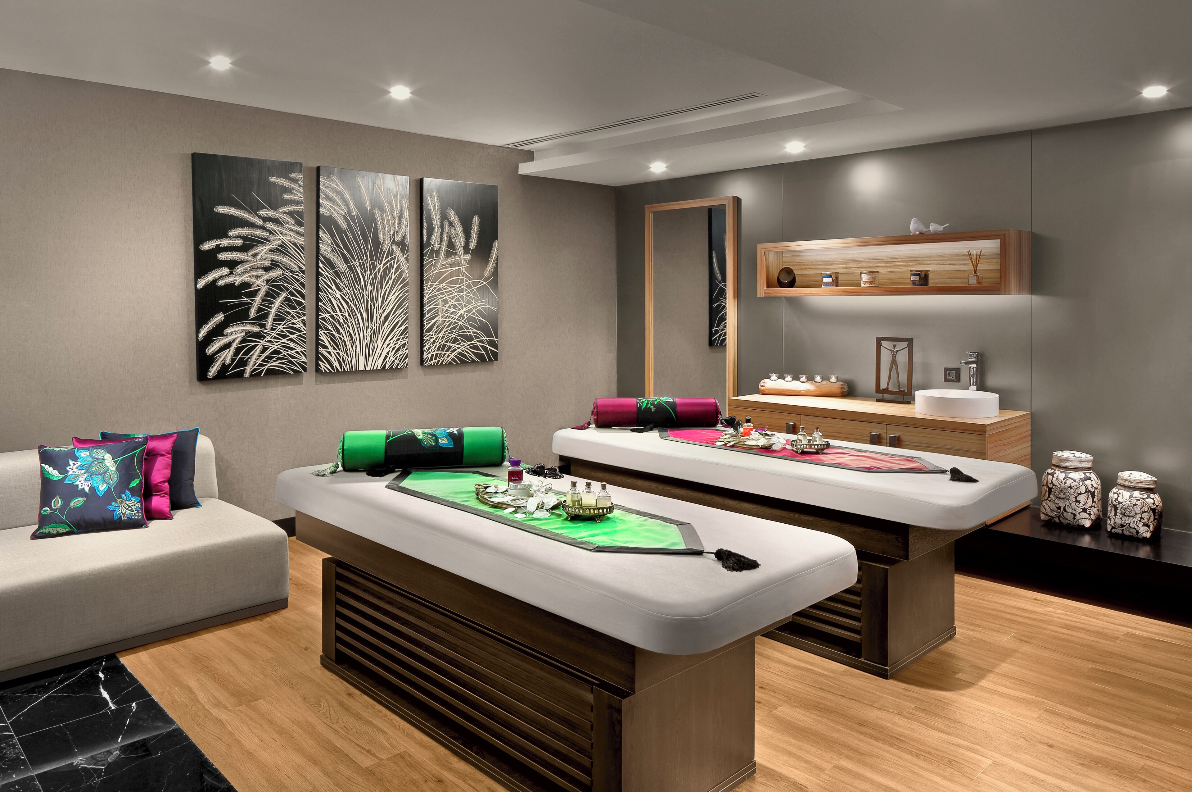 A spa room with a massage table, a sofa with cushions, a mirror, and wall-mounted shelves with decorative items.
