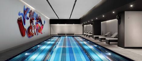 A modern indoor swimming pool with clear lanes, lounge chairs, and a large abstract art piece on the wall.