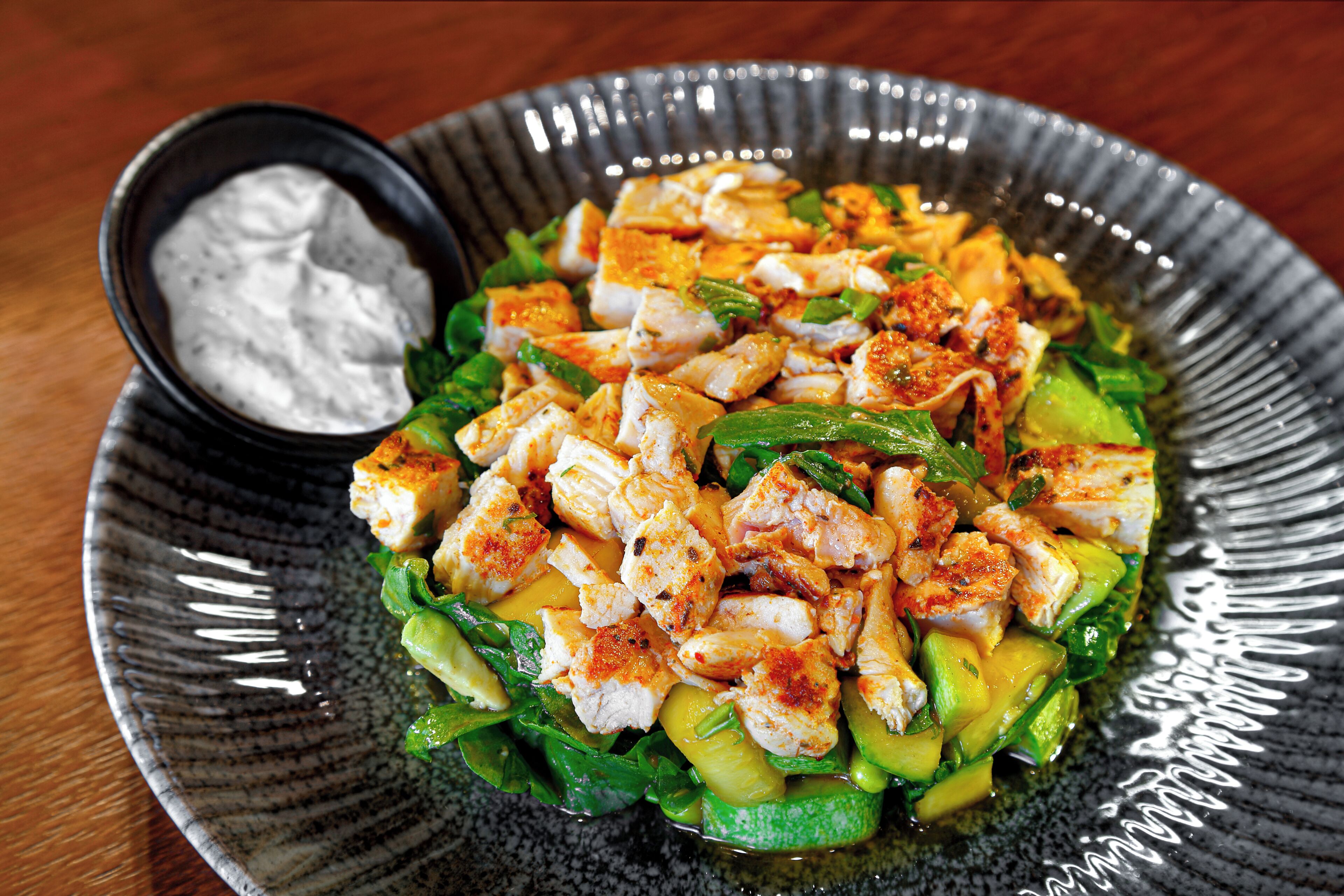 Un plato de comida con pollo, aguacate y verduras, además de un recipiente pequeño con salsa.