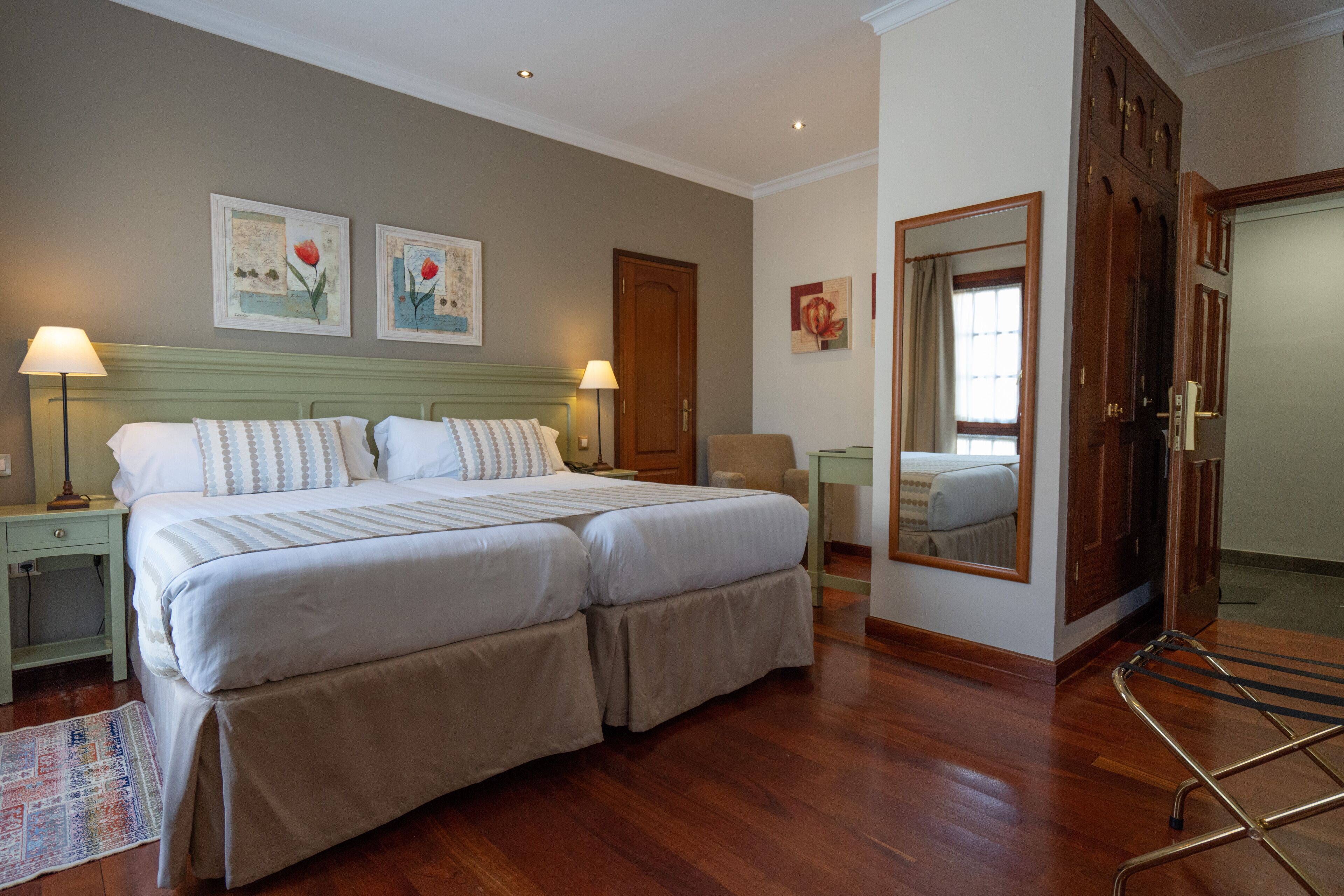 Una cama doble con dos mesitas de noche, una silla, un espejo y suelo de madera.