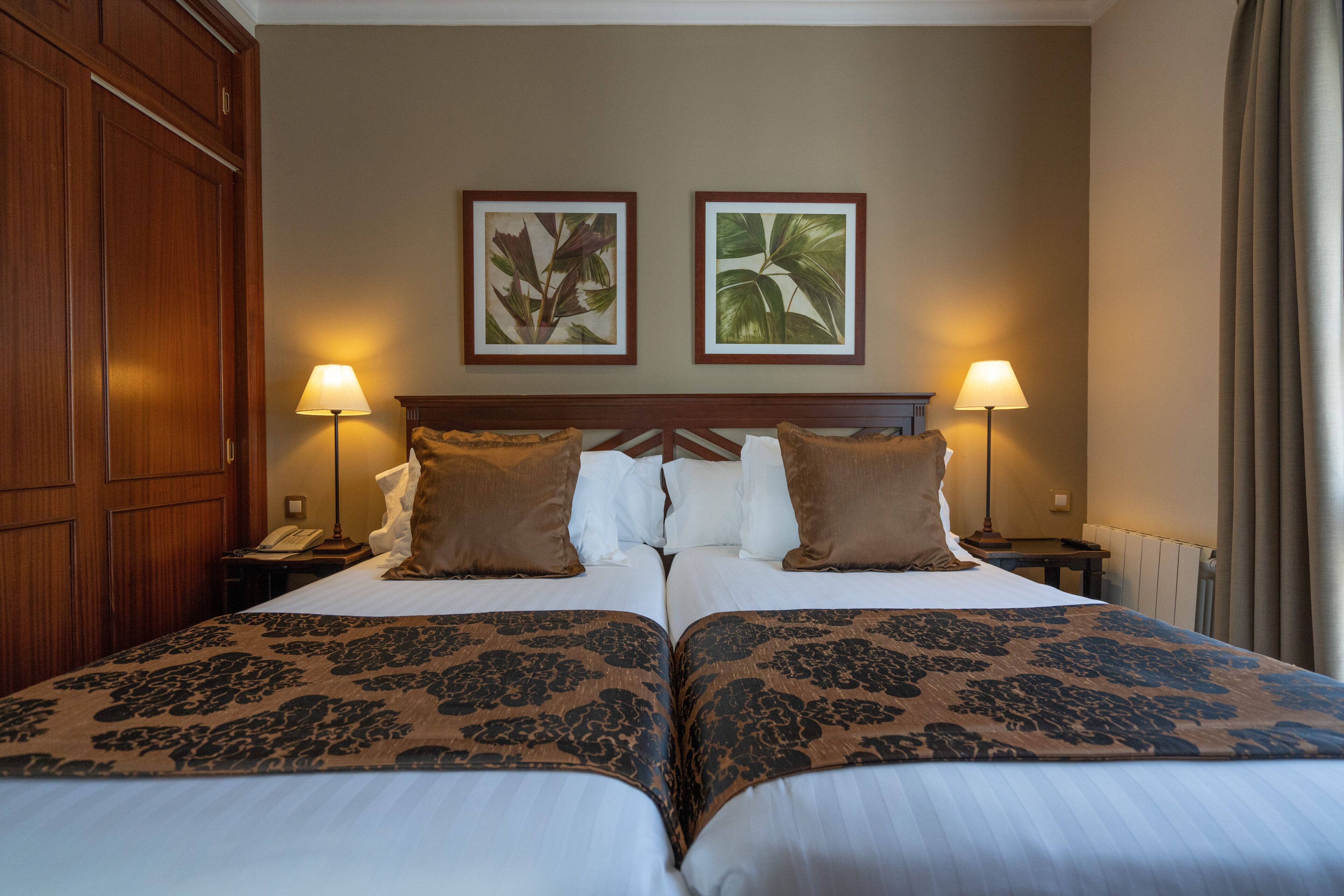 A hotel room with two beds, a wooden wardrobe, a nightstand with a lamp, and two framed botanical prints on the wall.