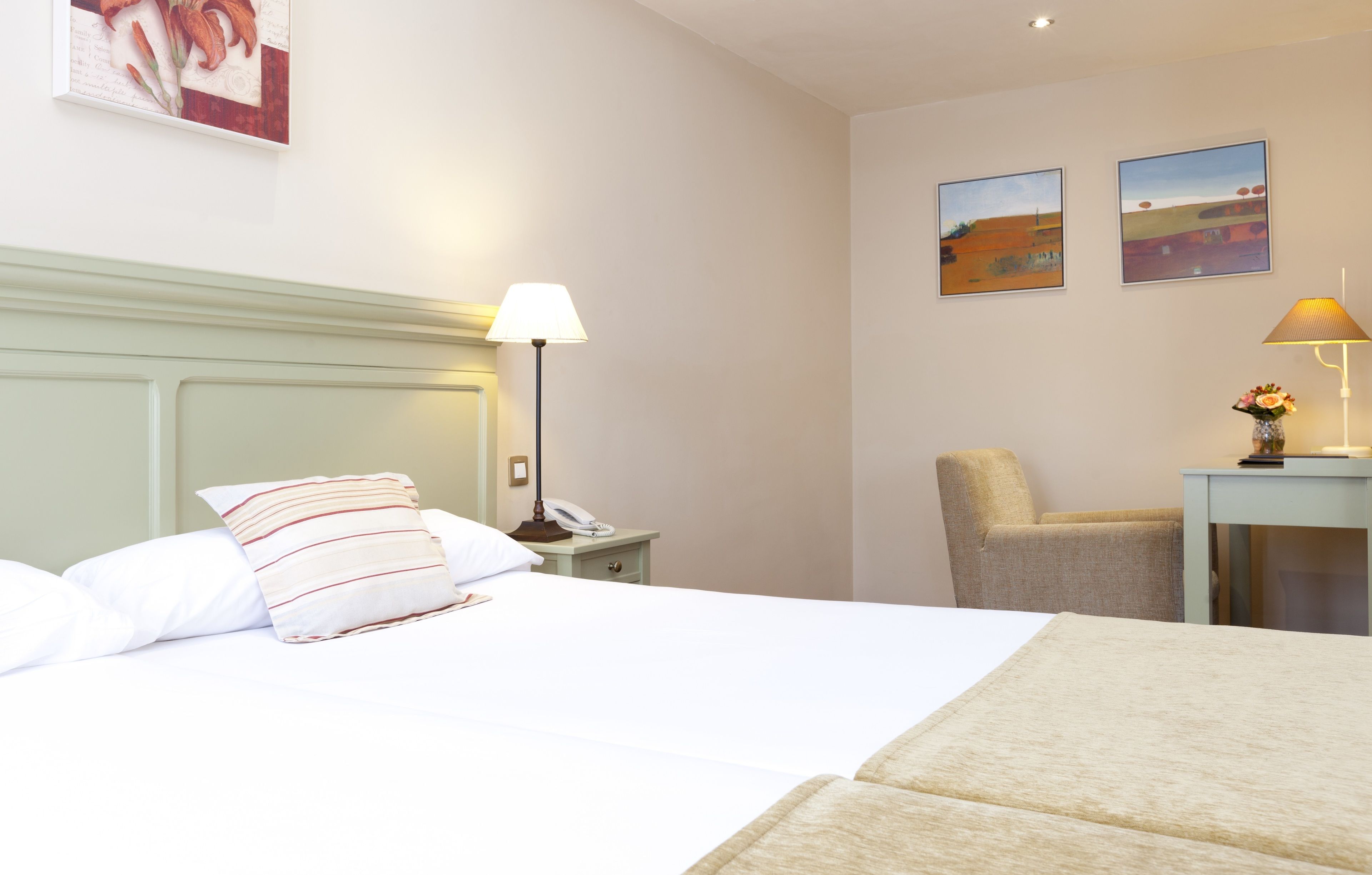 A hotel room with a bed, a nightstand with a lamp, a chair, and two wall-mounted paintings.