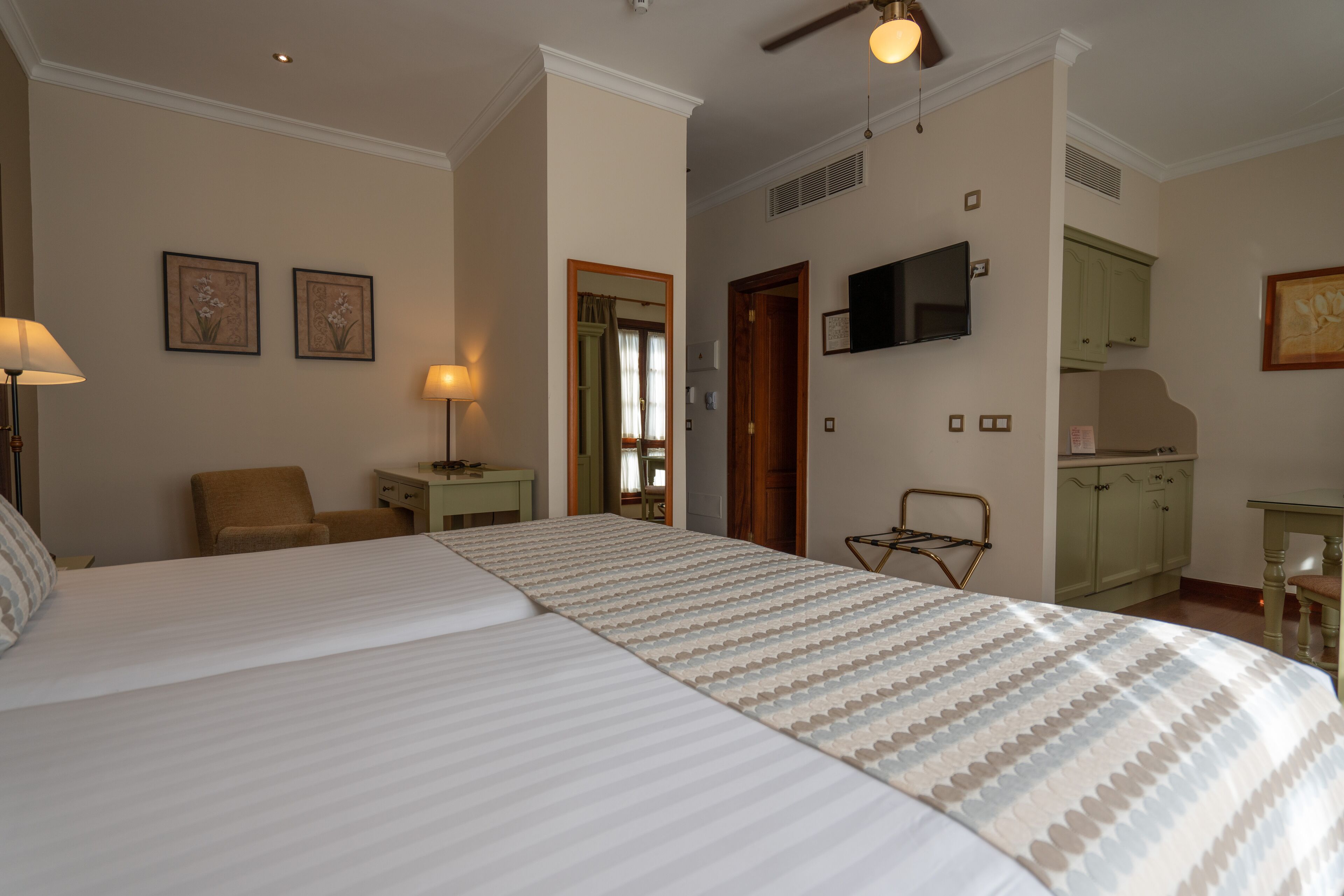 Habitación de hotel con una cama grande, un escritorio con lámpara, una silla, televisor montado en la pared y una zona de kitchenette.
