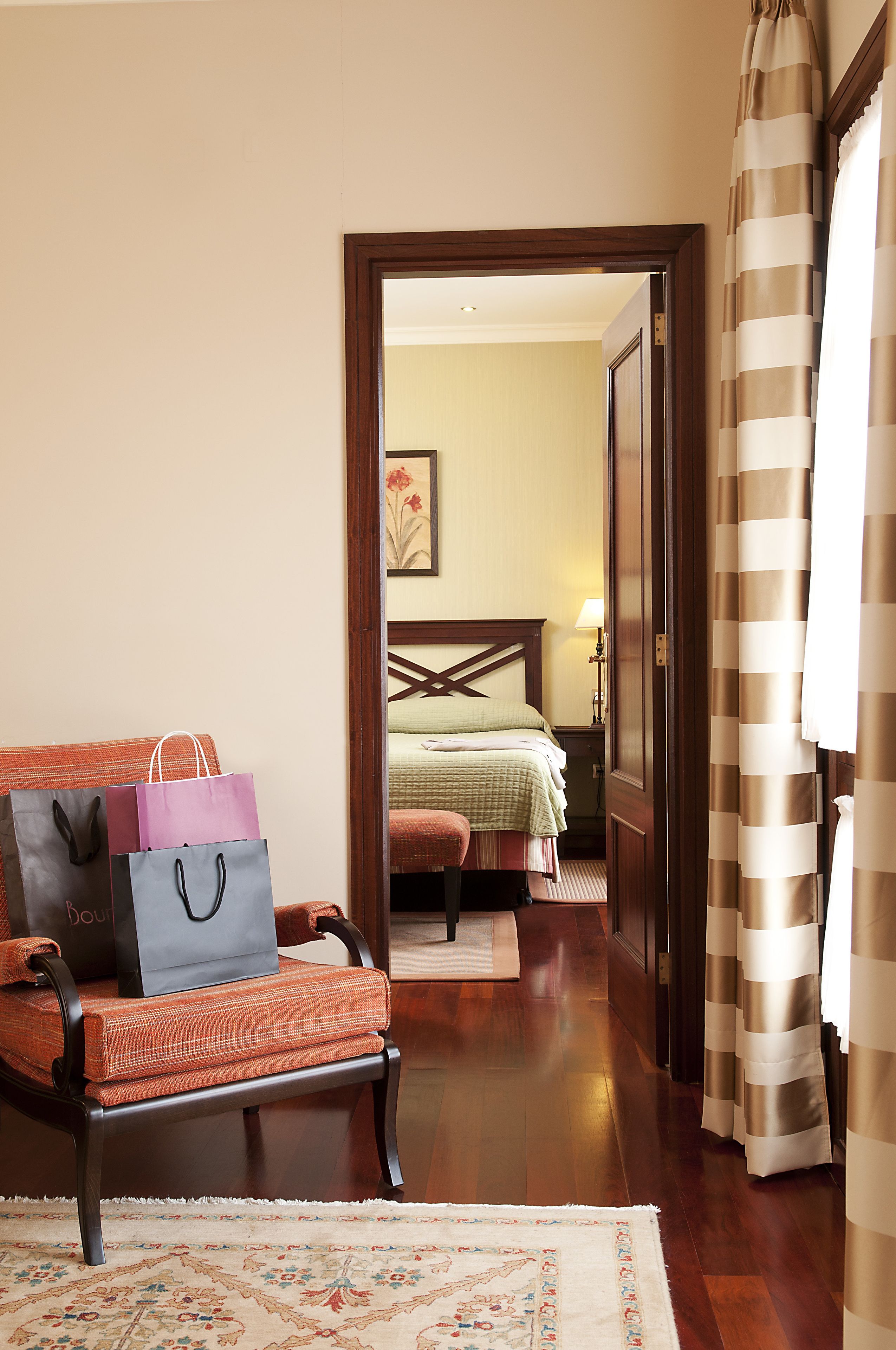A hotel room with a bed, a chair with shopping bags, and a view into the adjacent room.