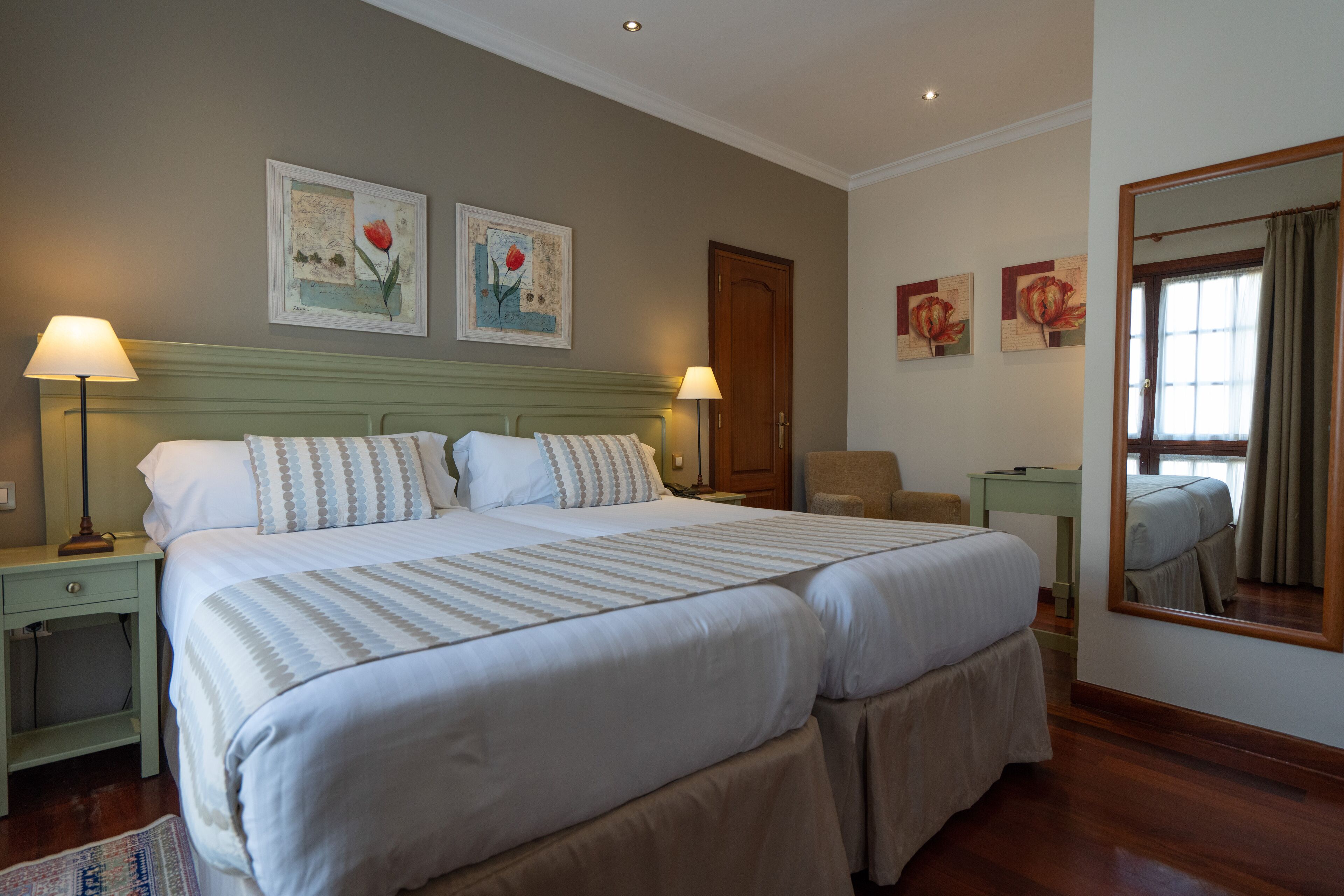 A double bed with a striped bedspread, two bedside lamps, a nightstand, a chair, a desk, and a mirror.