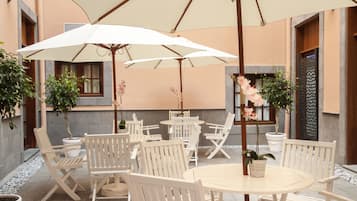 An outdoor seating area with white tables and chairs, surrounded by potted plants and umbrellas.