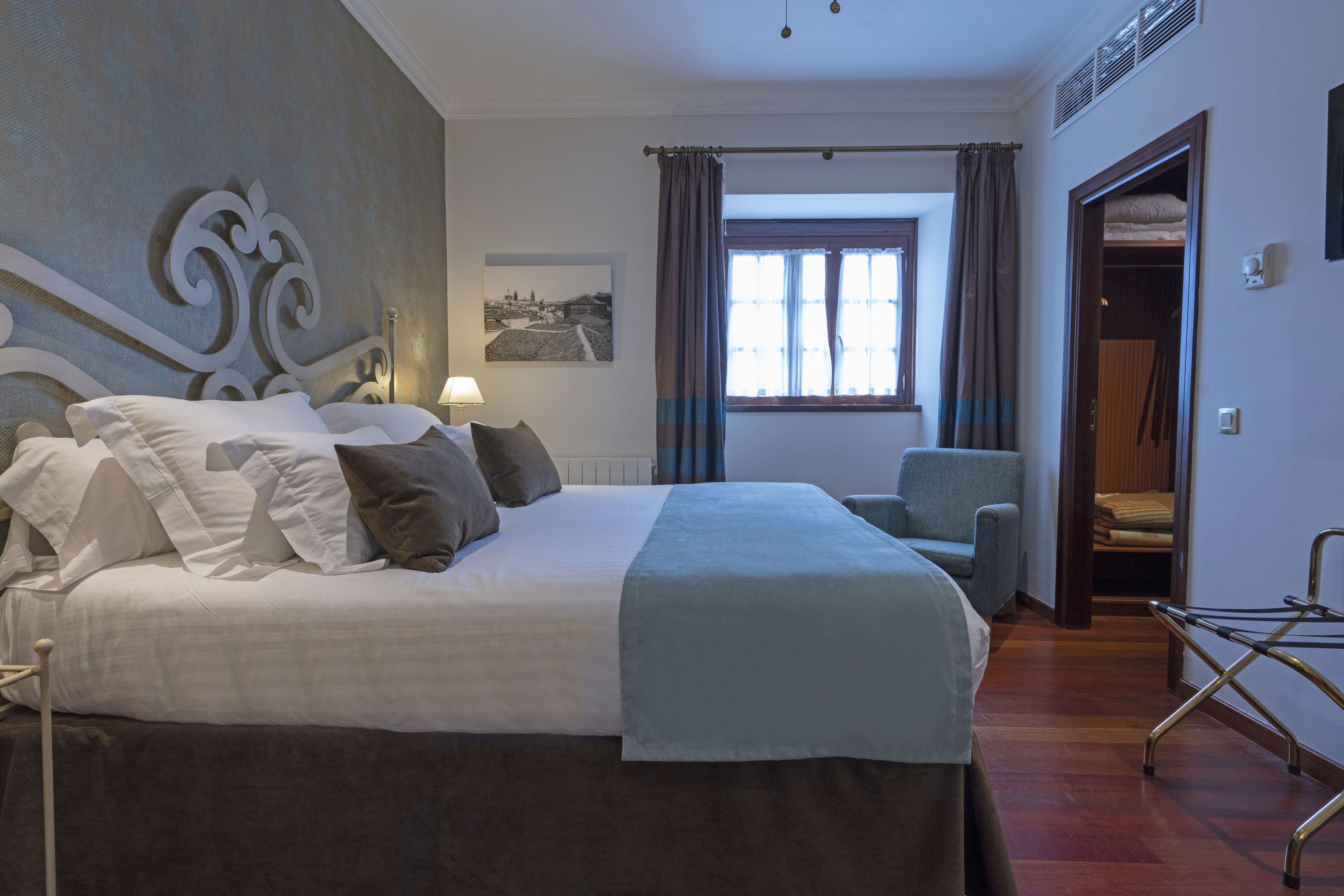 A hotel room with a large bed, a chair, a window with curtains, a picture on the wall, and a door leading to another room.