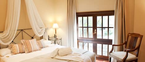 Un dormitorio con una cama grande, una silla de madera, un ventanal con cortinas y una mesita de noche con lámpara.