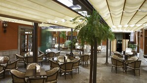 A spacious outdoor café area with multiple tables and chairs, shaded by large umbrellas, and surrounded by potted plants.