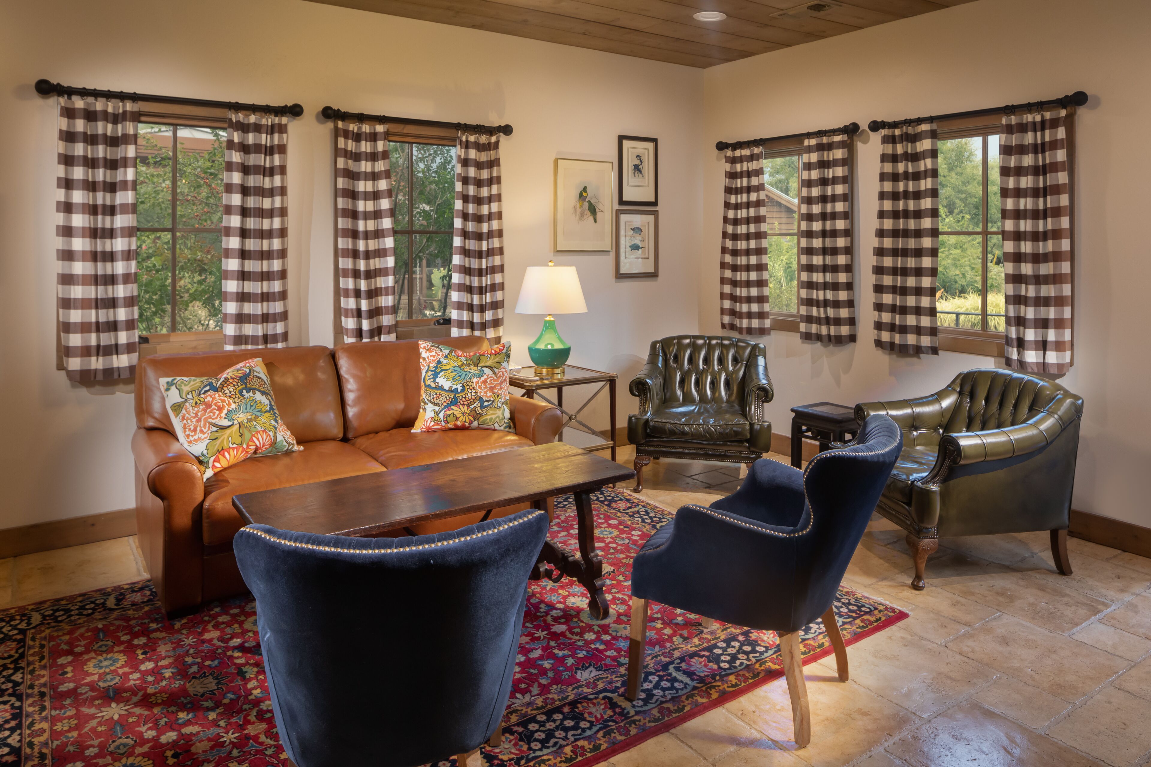 A living room with a brown leather sofa, a wooden coffee table, two blue armchairs, and a green armchair. There is a patterned rug on the floor, and the room has large windows with checkered curtains.