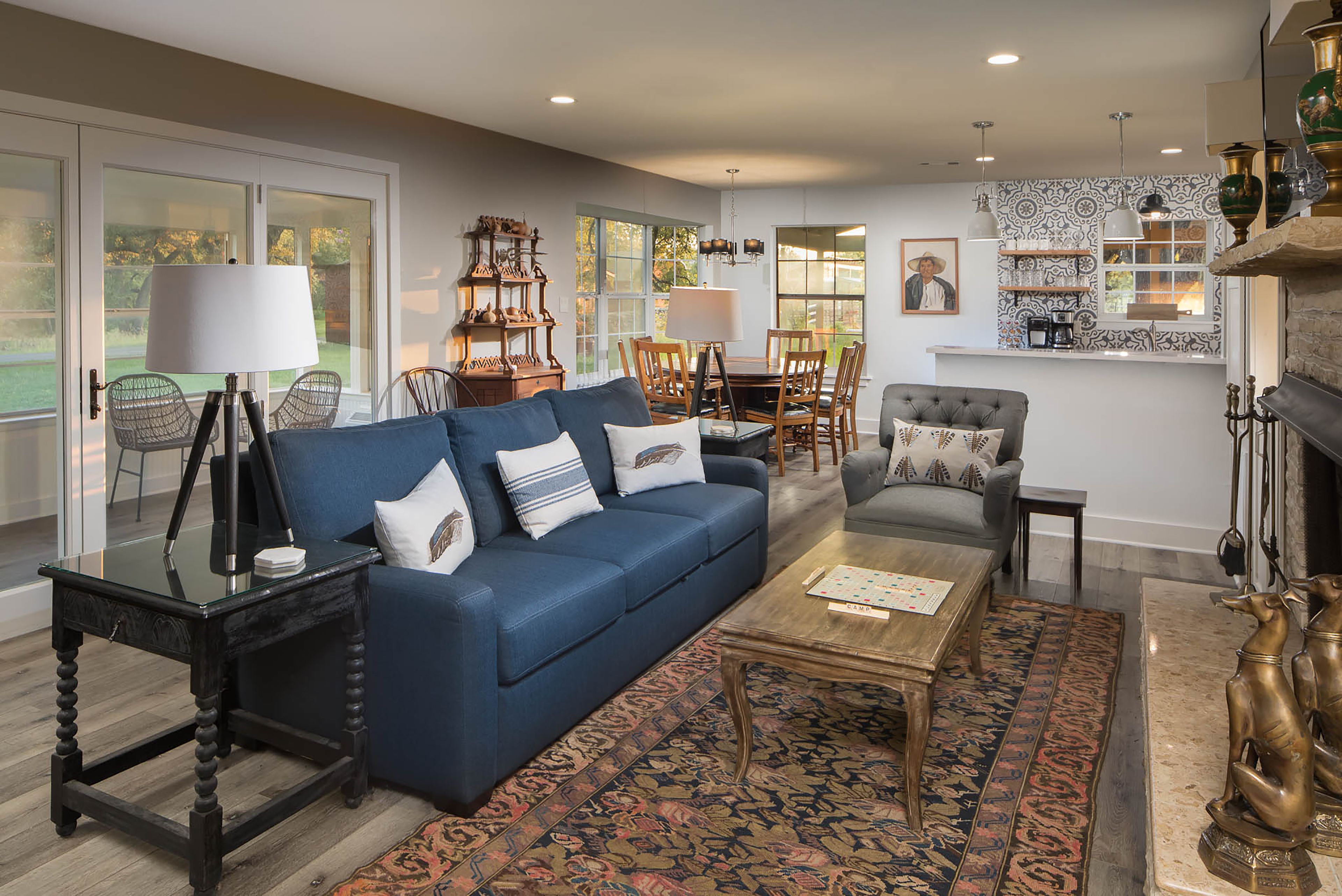 A spacious living room with a blue sofa, a coffee table, and a patterned rug. There is a dining area with a table and chairs, and a fireplace with decorative items.