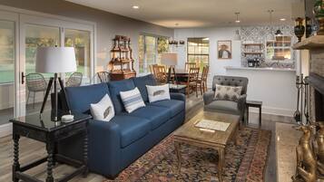 A spacious living room with a blue sofa, a coffee table, and a patterned rug. There is a dining area with a table and chairs, and a fireplace with decorative items.