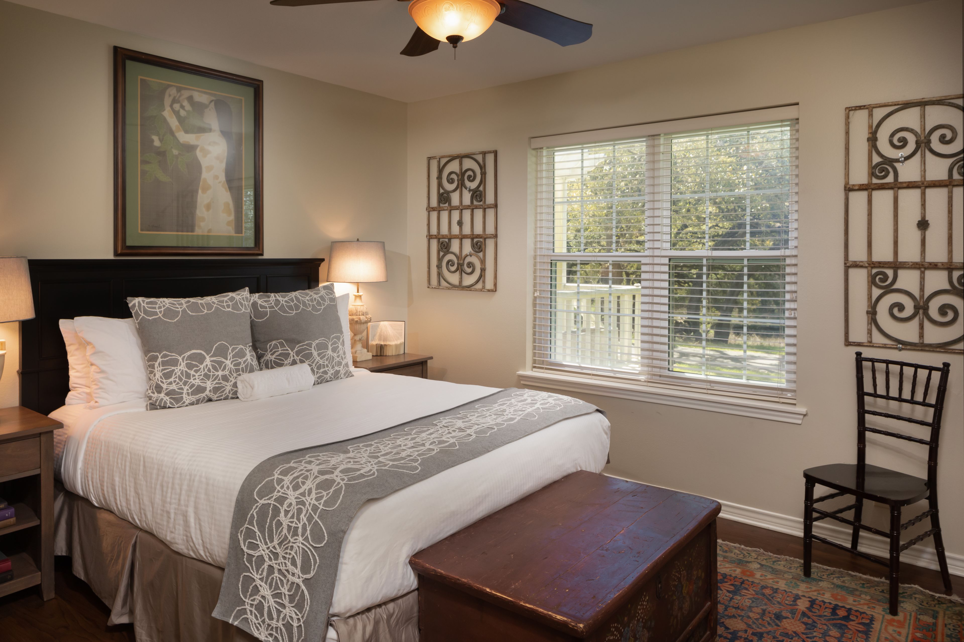 A neatly arranged bedroom with a large bed, bedside lamps, a window with blinds, decorative wall art, and a wooden bench.
