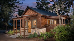 A rustic log cabin with a stone foundation, wooden porch, and a metal roof. - Camp Lucy (Dripping Springs)