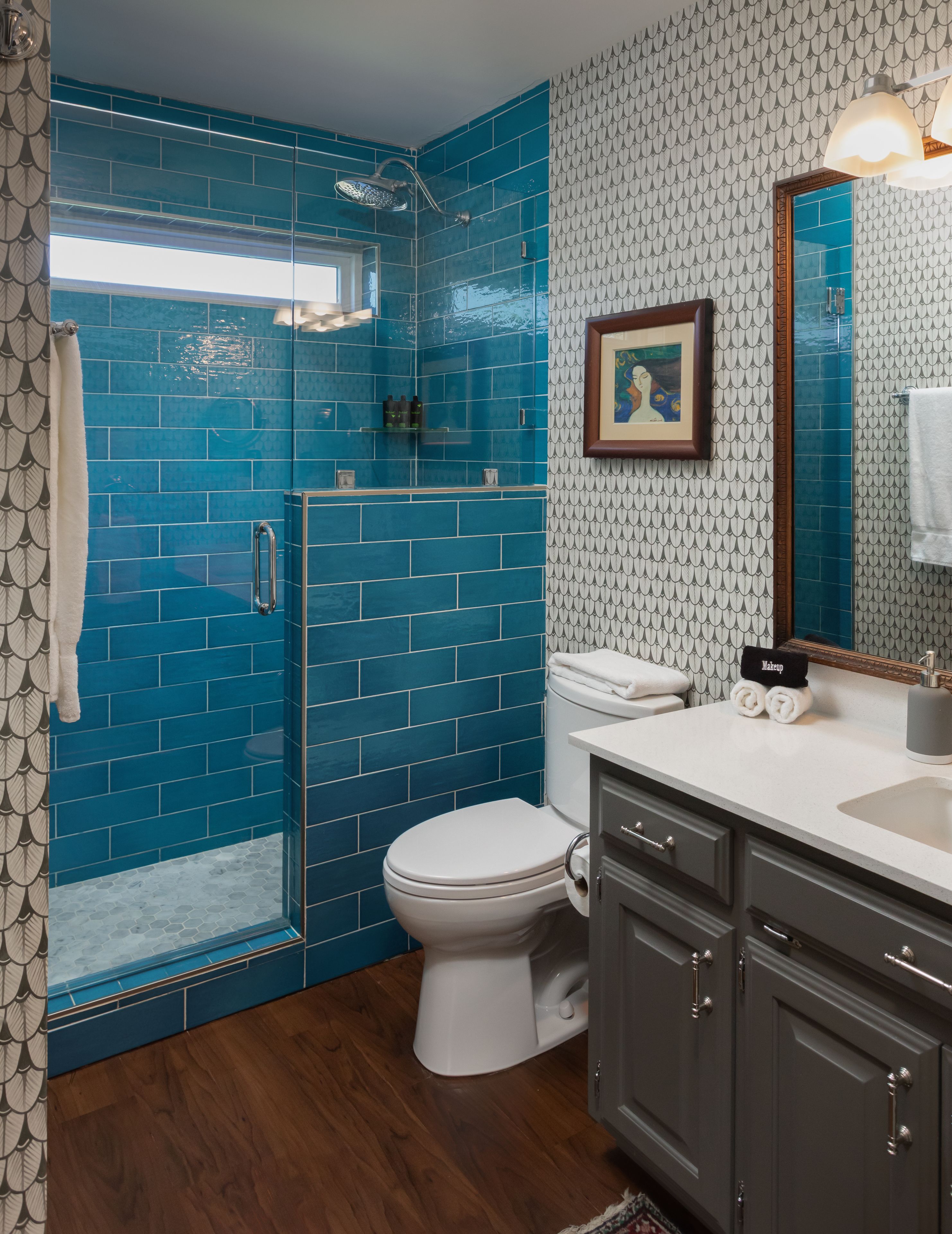 A bathroom with a blue-tiled shower, a white toilet, and a vanity with a mirror.