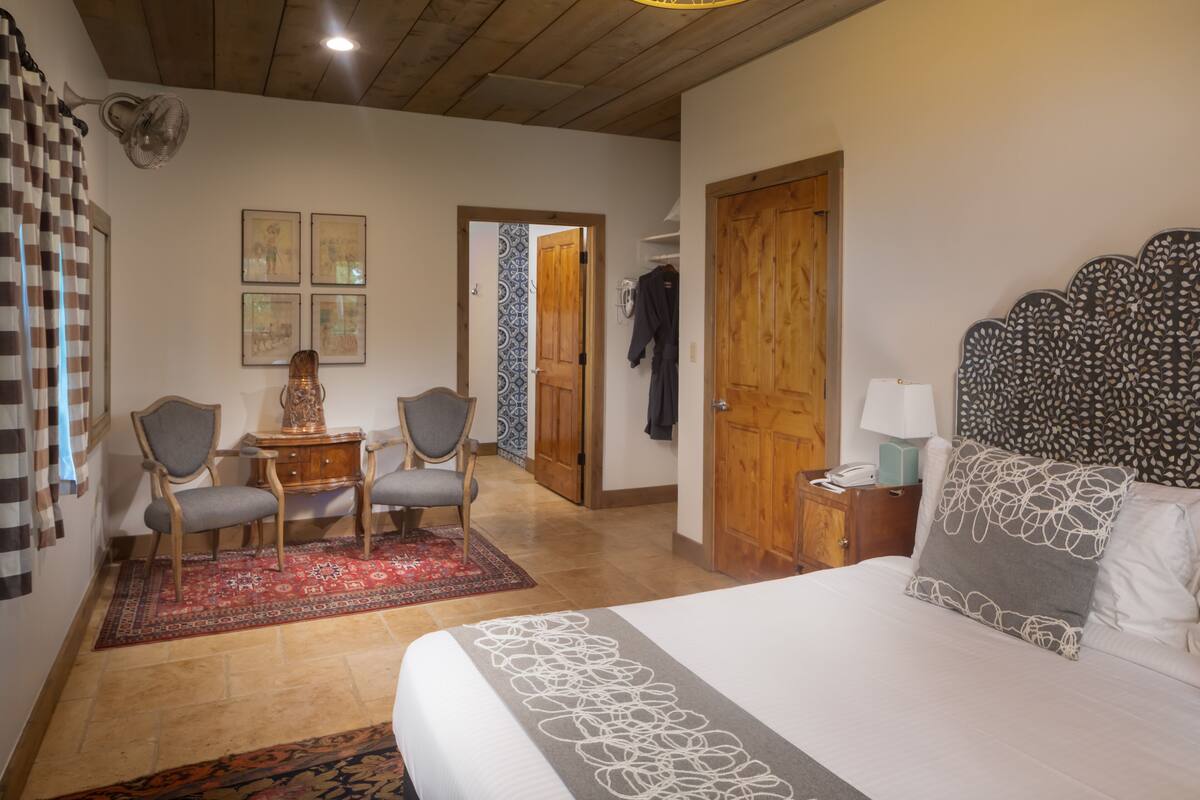 Ein Hotelzimmer mit einem Bett, zwei Stühlen, einem kleinen Tisch, einem roten Teppich und einer Holztür.