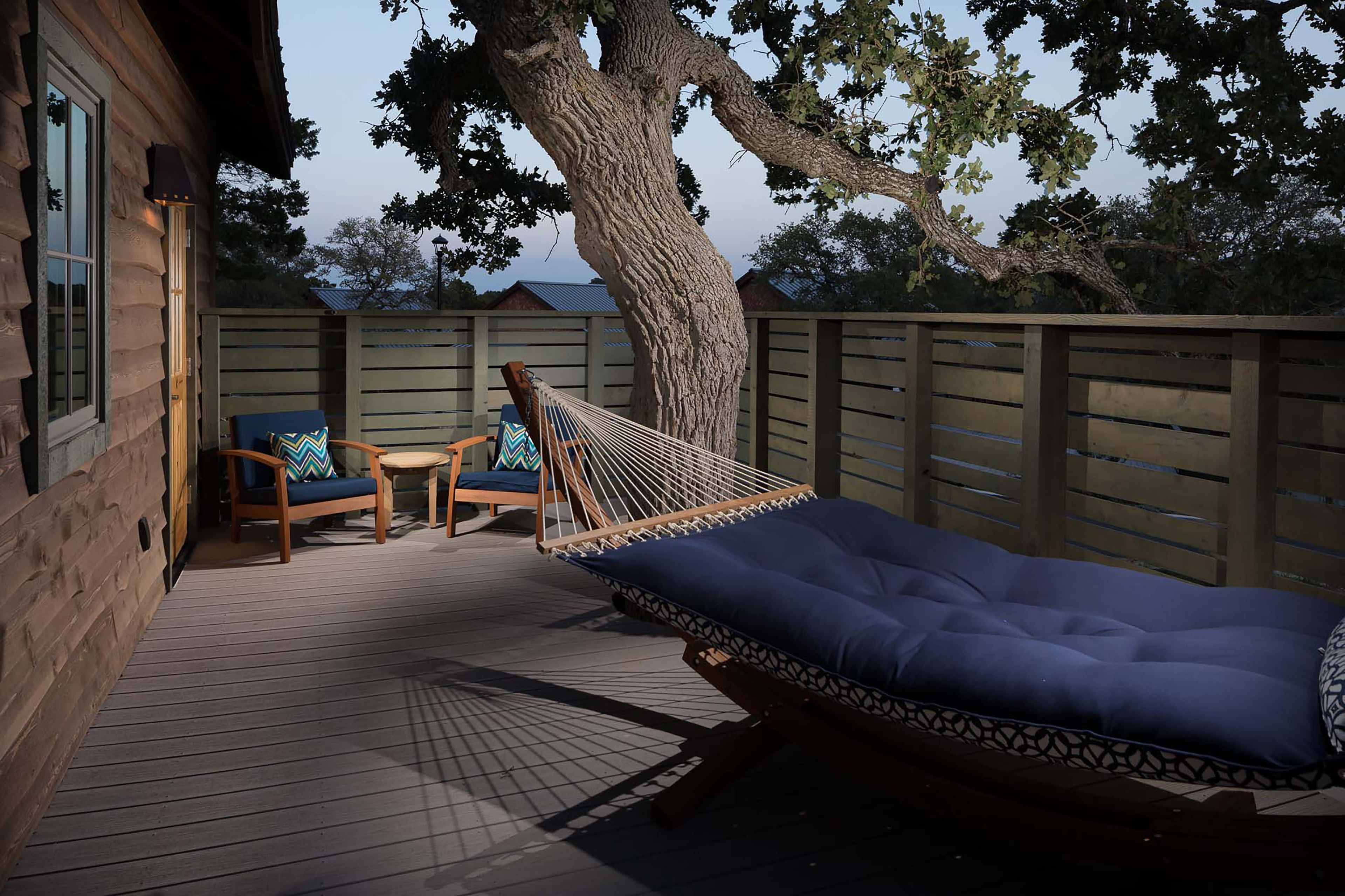 A wooden deck with a hammock, two chairs, and a view of a tree and building.