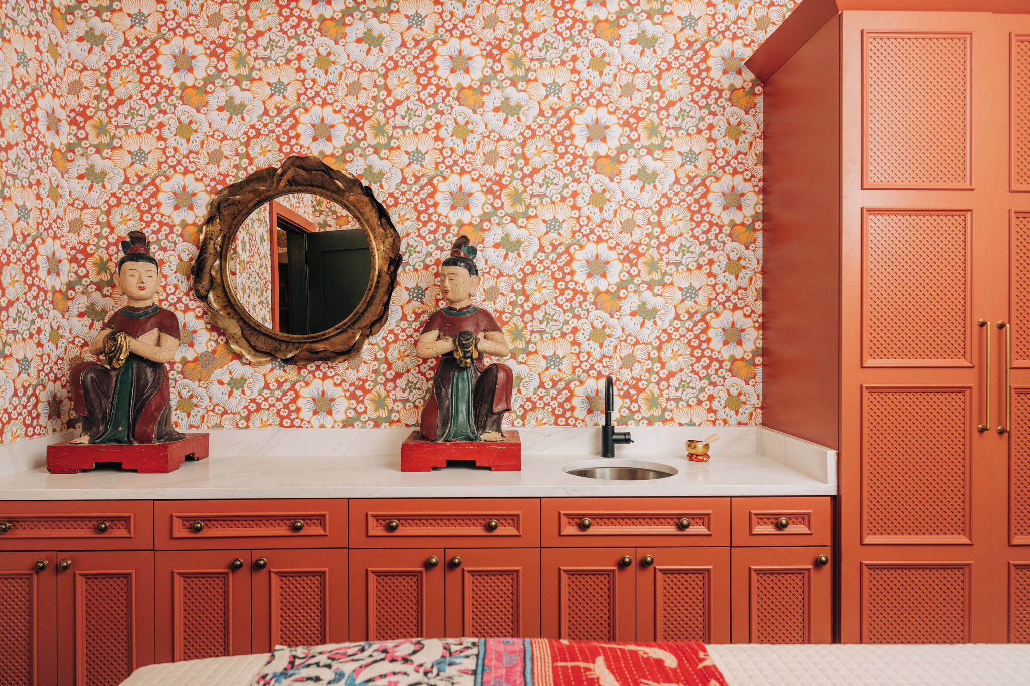 Une salle de bain avec un papier peint à motifs floraux, deux statues posées sur le meuble-lavabo, un miroir et un meuble rouge.