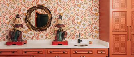 Une salle de bain avec un papier peint à motifs floraux, deux statues posées sur le meuble-lavabo, un miroir et un meuble rouge.