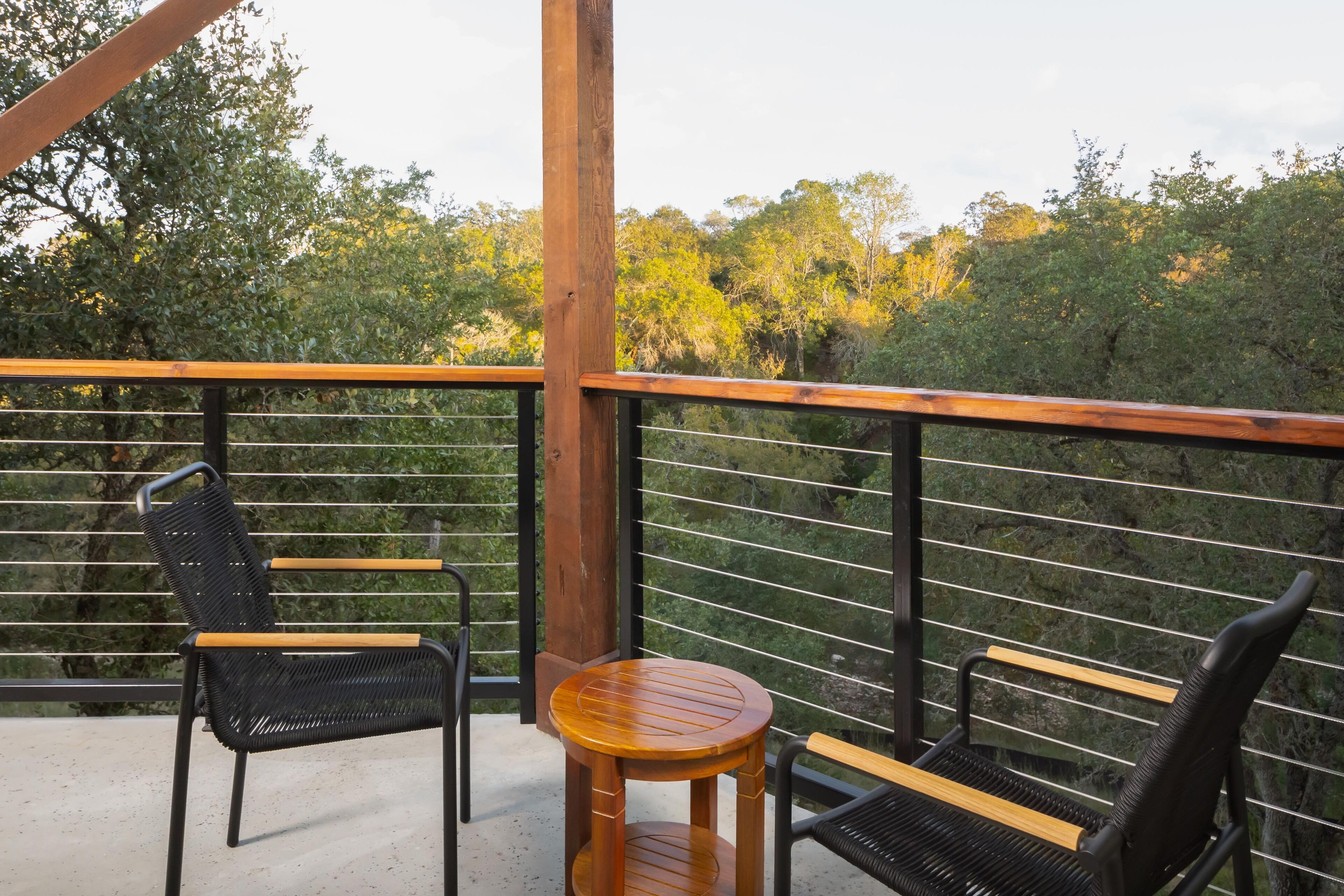 A balcony with two black wicker chairs and a wooden table, overlooking a forested area.