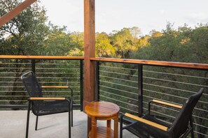 A balcony with two black wicker chairs and a wooden table, overlooking a forested area. - Camp Lucy (Dripping Springs)