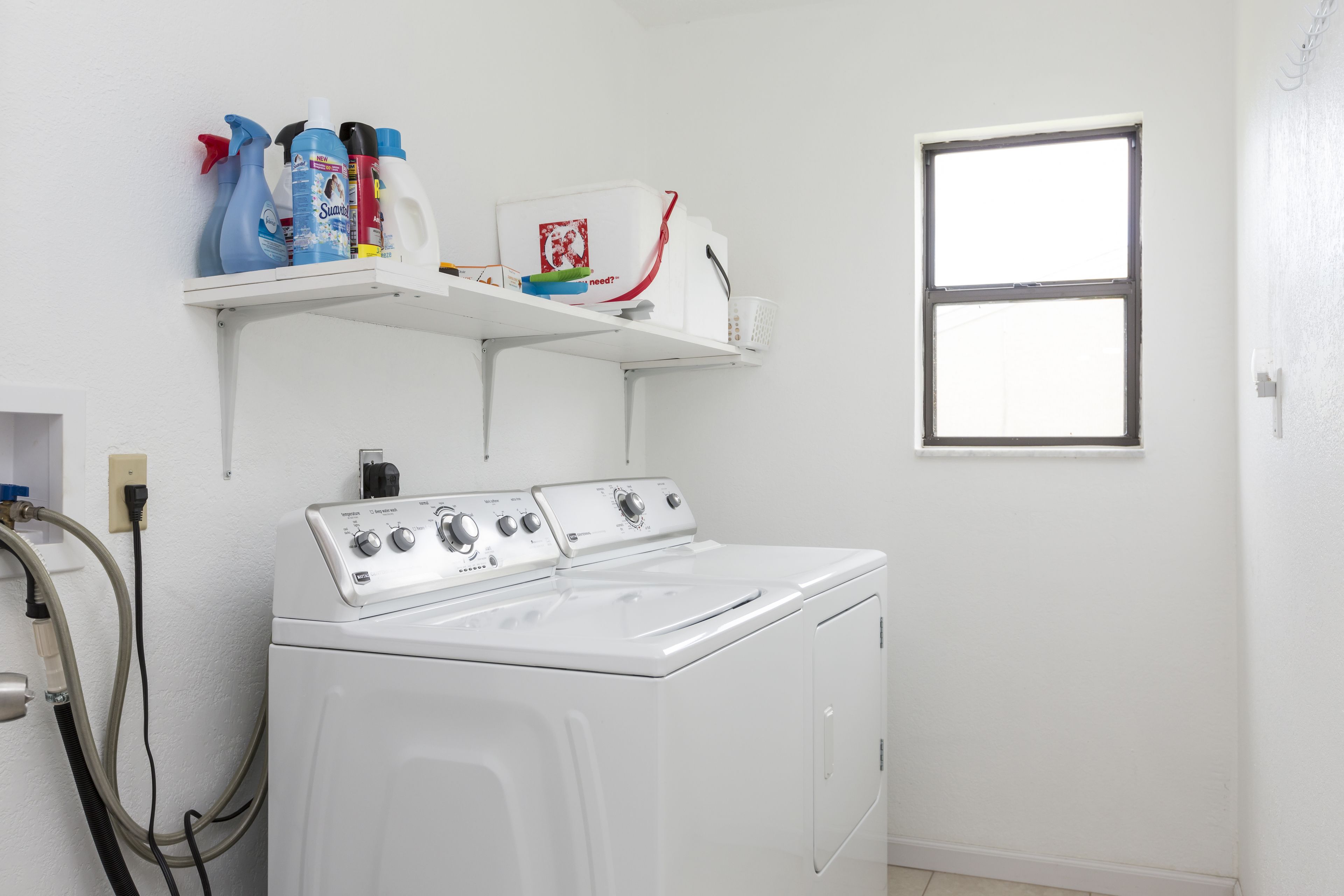 A laundry room with a front-loading washer and dryer, a shelf with cleaning supplies, and a window.