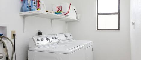 A laundry room with a front-loading washer and dryer, a shelf with cleaning supplies, and a window.