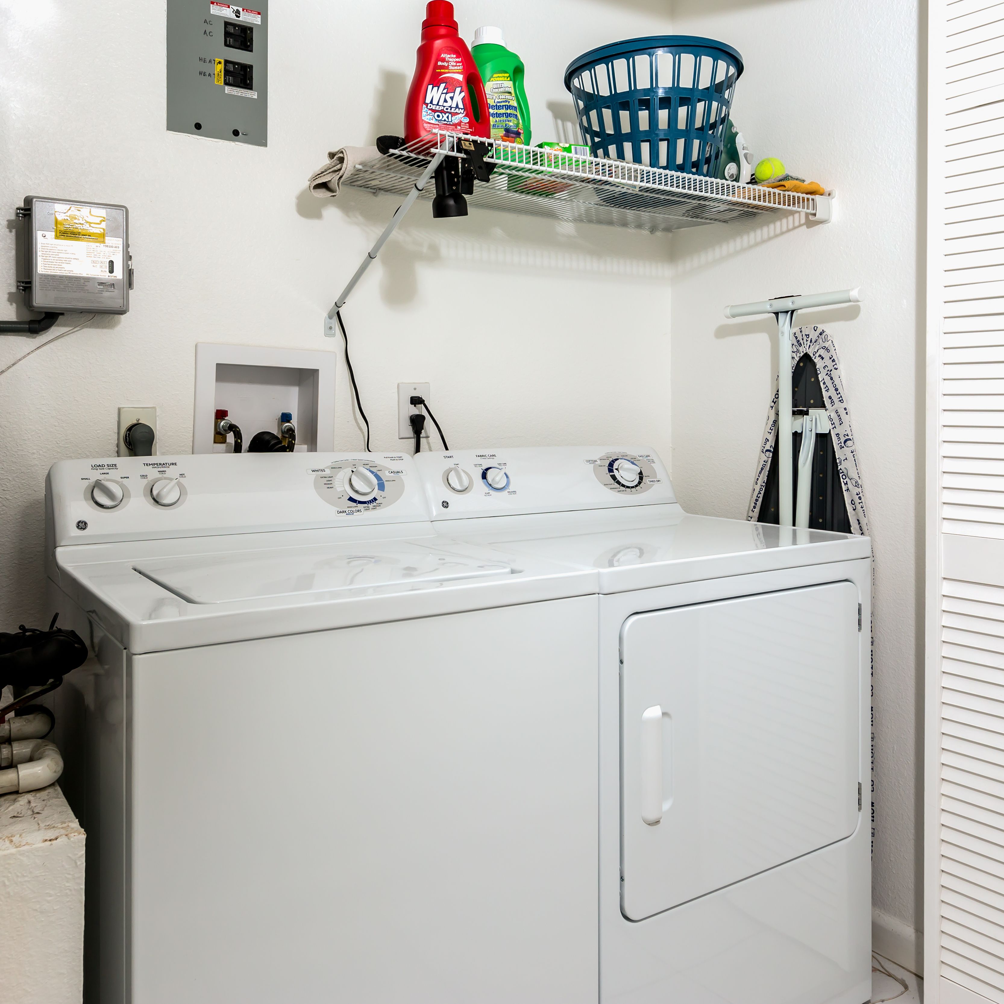 A front-loading washer and dryer in a laundry room with a shelf above containing cleaning supplies.