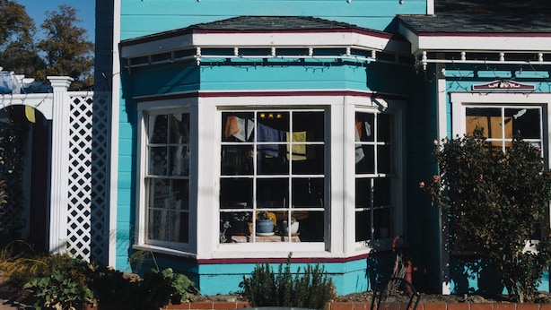 Une maison à deux étages avec un toit rose et un revêtement extérieur turquoise, dotée d’une baie vitrée et d’un petit patio avec une table et des chaises.