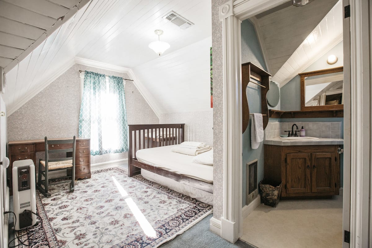 A bedroom with a bed, a desk, a chair, a radiator, and a bathroom with a sink and mirror.