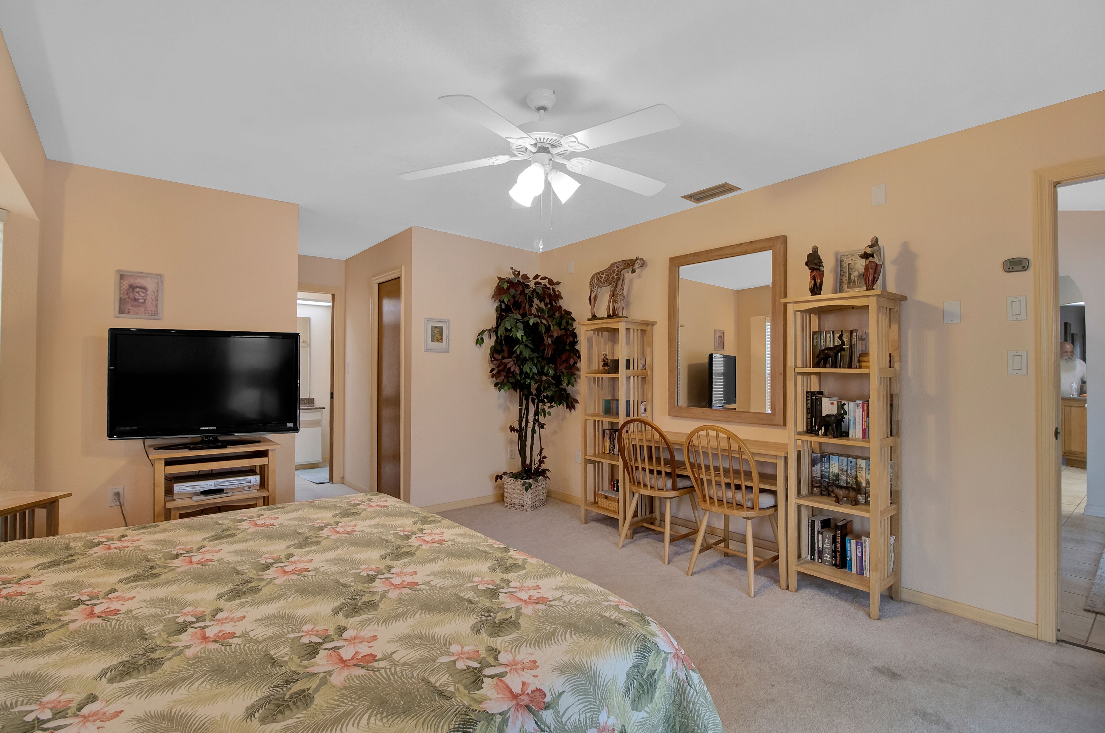 A bedroom with a bed, a TV, a desk with a chair, a bookshelf, and a ceiling fan.