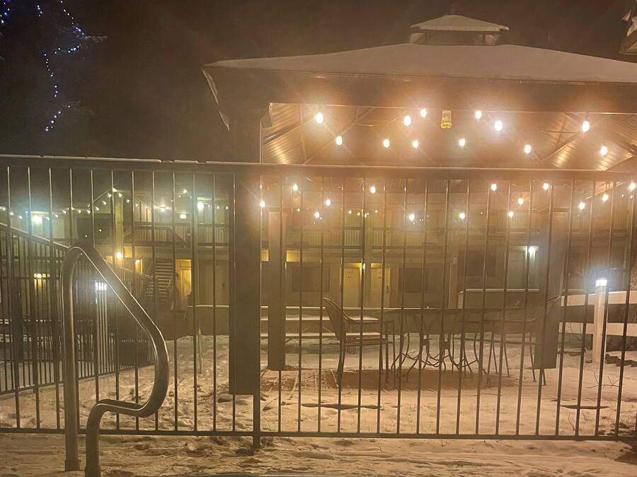 A snow-covered outdoor area with a hot tub, metal railing, and a building with string lights.