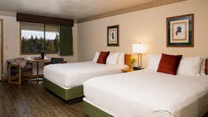 A hotel room with two beds, a small table with a chair, a window with a view of trees, and framed artwork on the walls. - Timbers Motel (Bigfork)