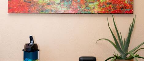 A painting of a forest scene on a wall, a coffee maker, a tray with mugs, a book, and a potted plant on a counter.