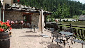A stone building with a tiled roof, outdoor seating with metal chairs and tables, and a view of a forested area in the background. - Hotel Banchetta (Sestriere)