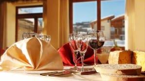 A table set with wine glasses, a cheese wheel, and bread, with a view of a snowy landscape through a window. - Hotel Banchetta (Sestriere)