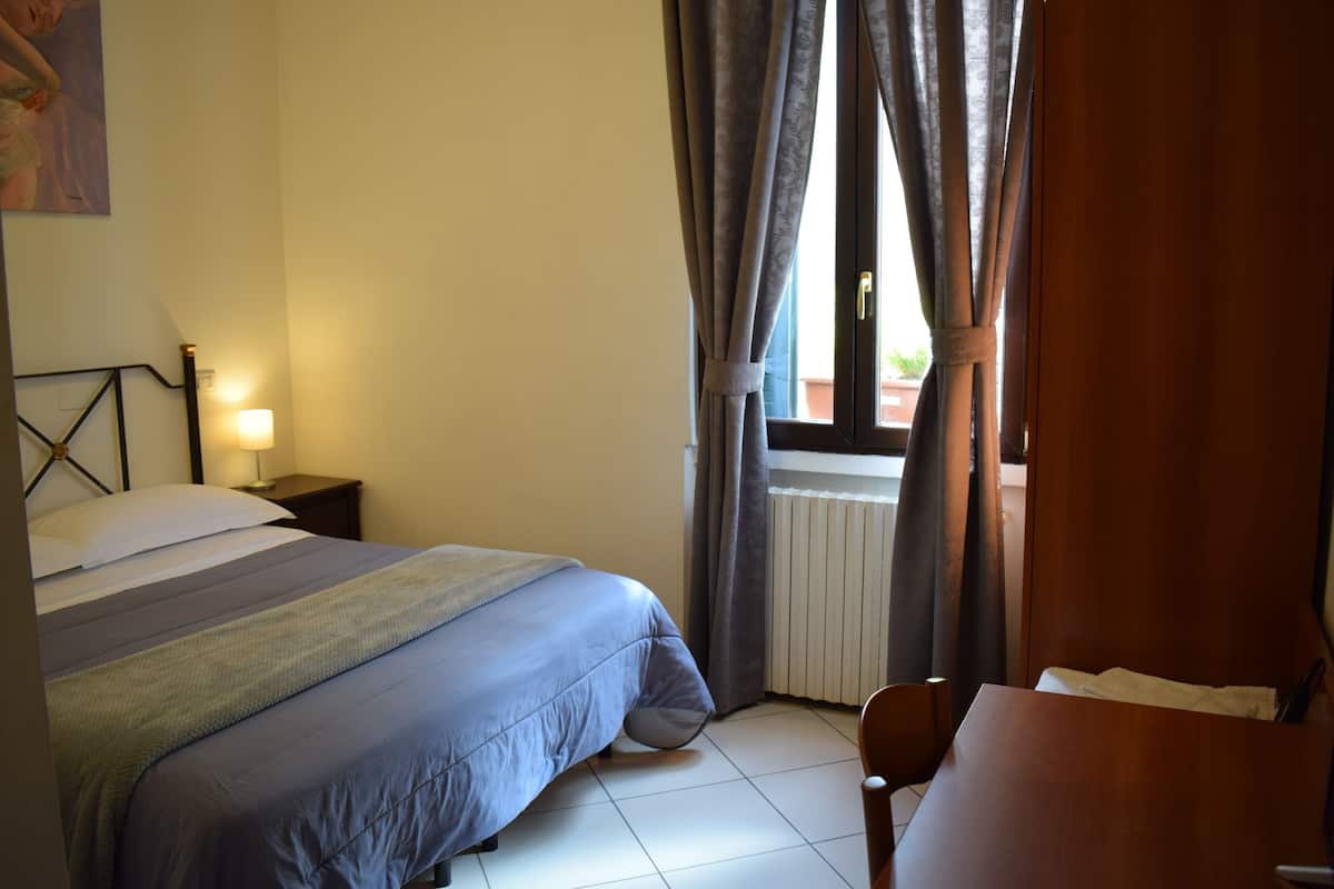 A bedroom with a bed, bedside table, lamp, and a view of the outdoors through curtains.