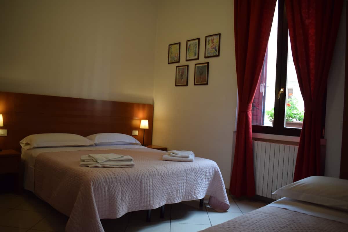 A hotel room with a double bed, a nightstand, a window with red curtains, and wall-mounted artwork.