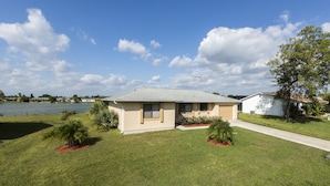 A single-story house with a flat roof, a front yard with small trees, and a view of a lake. - Lilsis by the Lake - Vacation Home (Port Charlotte)